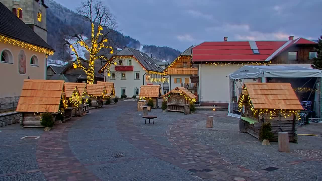A town square at dusk, decorated with small wooden huts strung with lights, a large tree also lit up with lights, and buildings with red roofs.