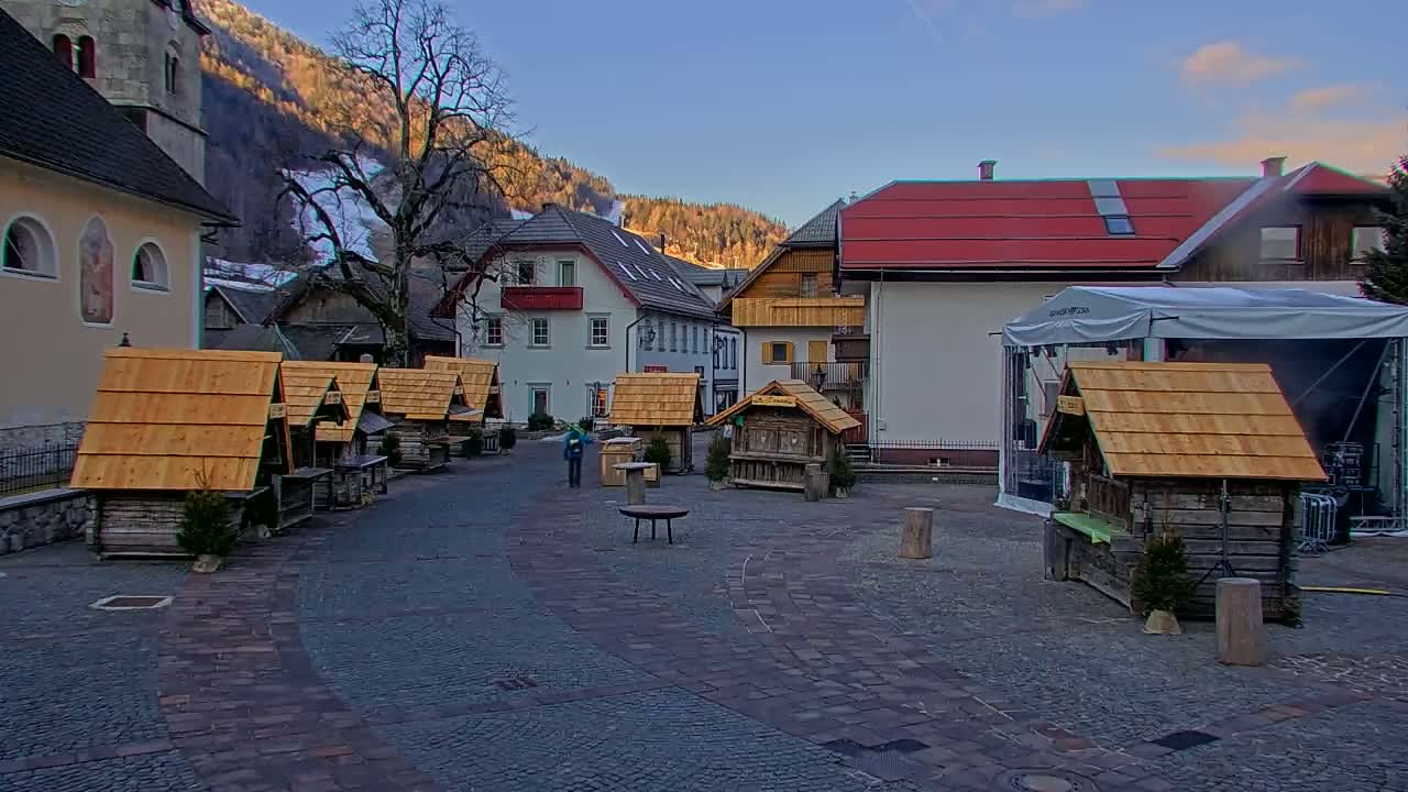A cobblestone town square, surrounded by buildings and small wooden stalls with peaked roofs, sits beneath a clear sky in a snow-dusted mountain village.