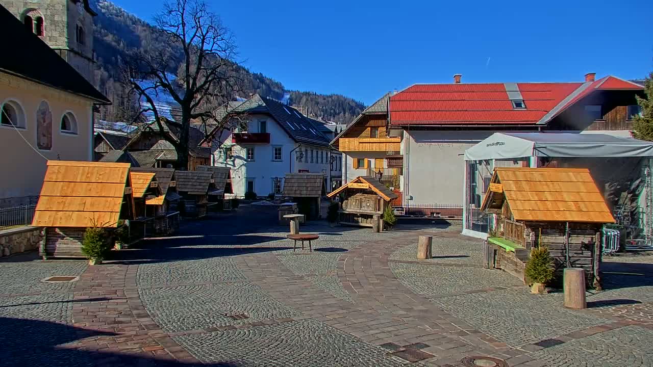 A sunny day reveals a cobblestone plaza with several small wooden structures, situated amongst buildings in a village nestled against snow-capped mountains.