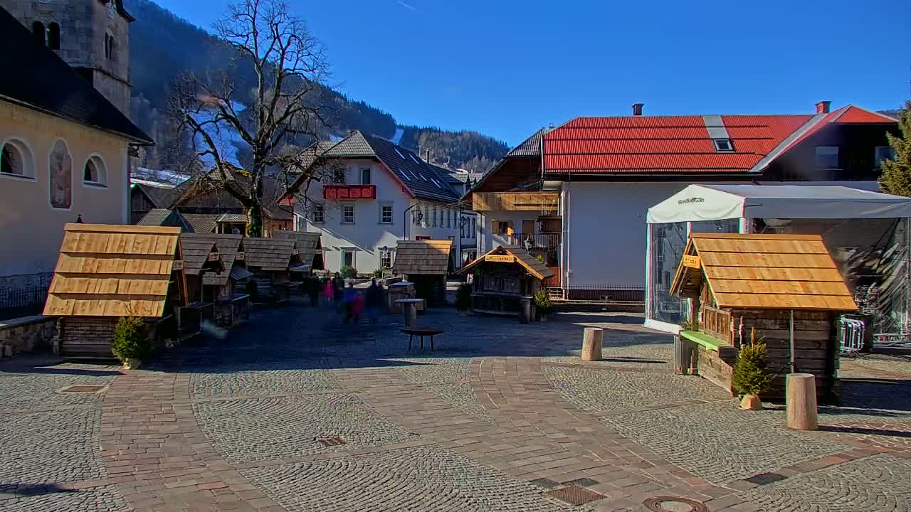 A cobblestone town square with several small wooden structures, surrounded by buildings, under a clear blue sky.
