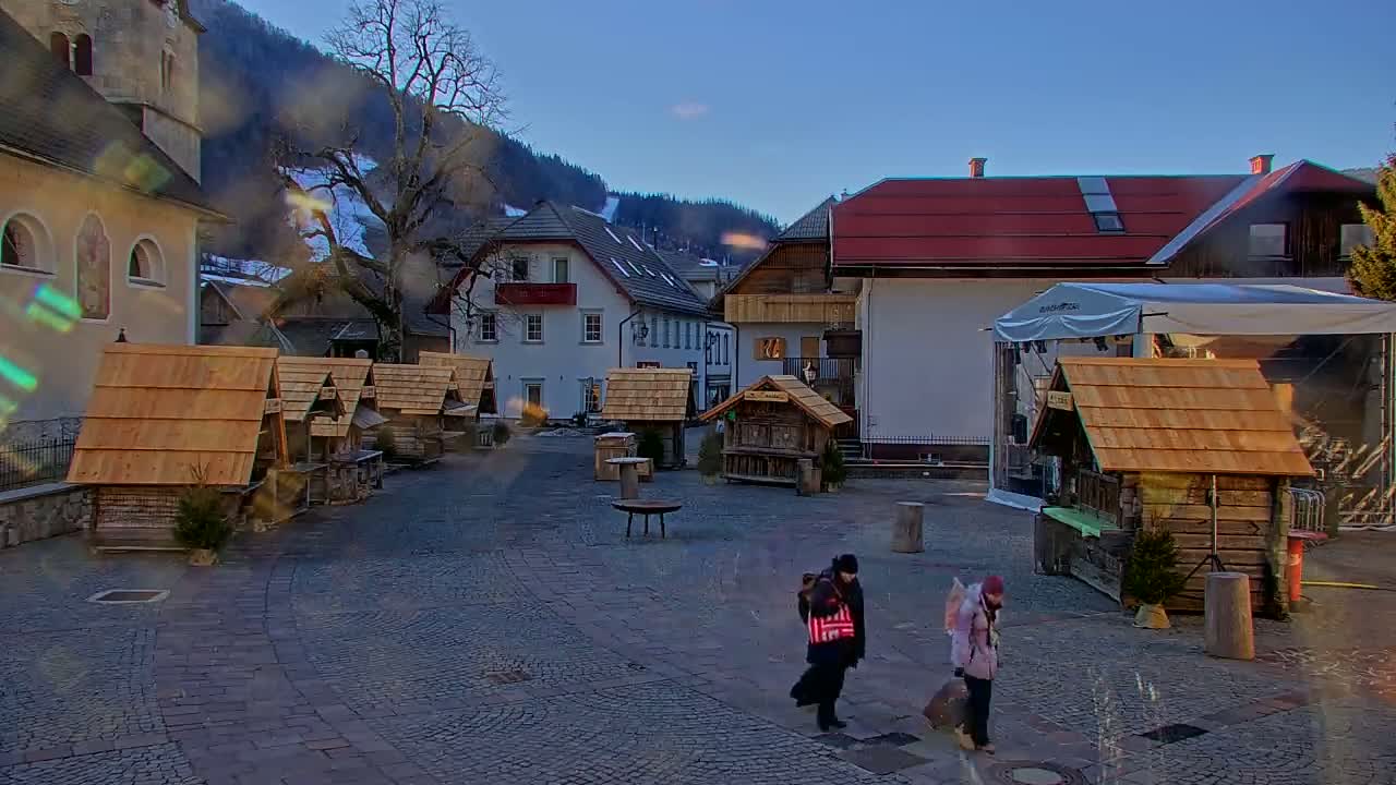 A cobblestone town square with several small wooden market stalls, buildings, and two people walking, under a clear, bright sky with snow visible on the distant mountains.