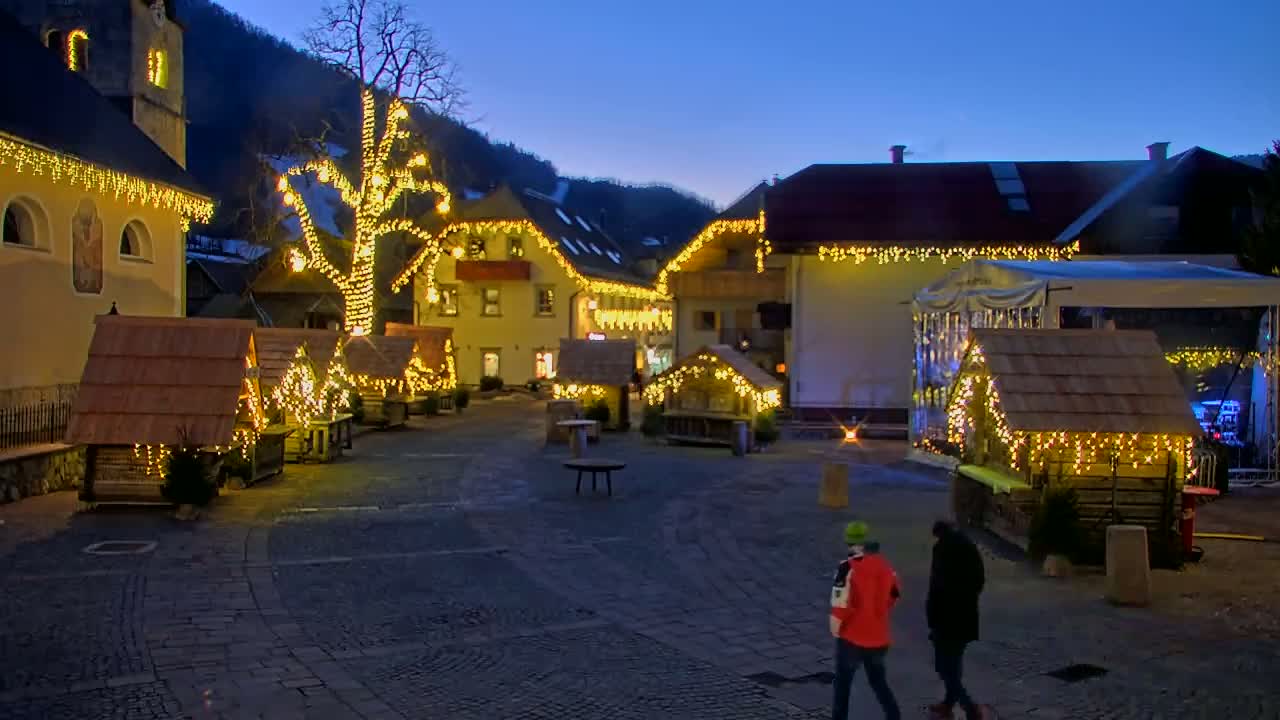 A town square at twilight is decorated with small wooden huts and string lights, two people walk through the square.