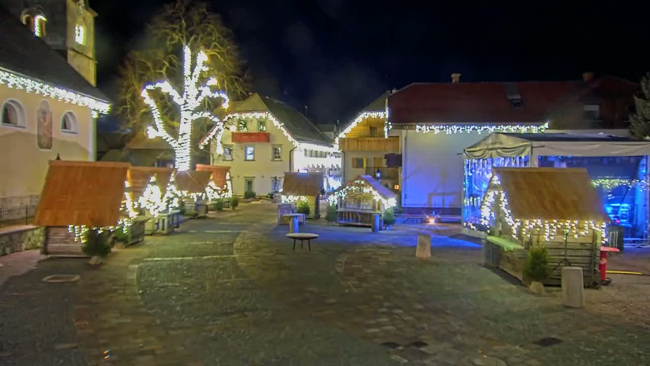 A town square at night is illuminated with strings of lights adorning buildings and small wooden structures, a large tree in the center is also decorated with lights.