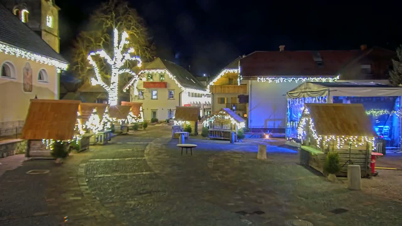A nighttime scene of a town square decorated with Christmas lights, featuring small wooden huts and a large building illuminated behind them.