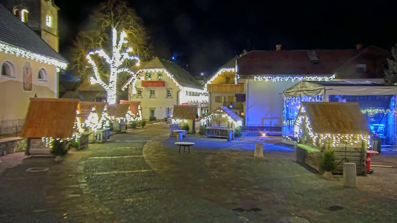 A nighttime scene of a town square decorated with Christmas lights and small wooden huts, illuminated against a dark sky.