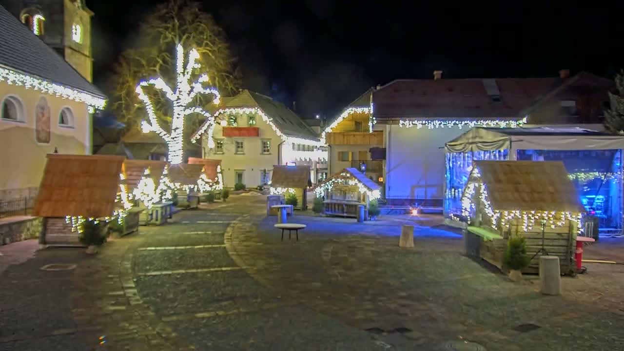 A nighttime scene of a town square decorated with Christmas lights, featuring several small wooden huts and a large tree also adorned with lights.