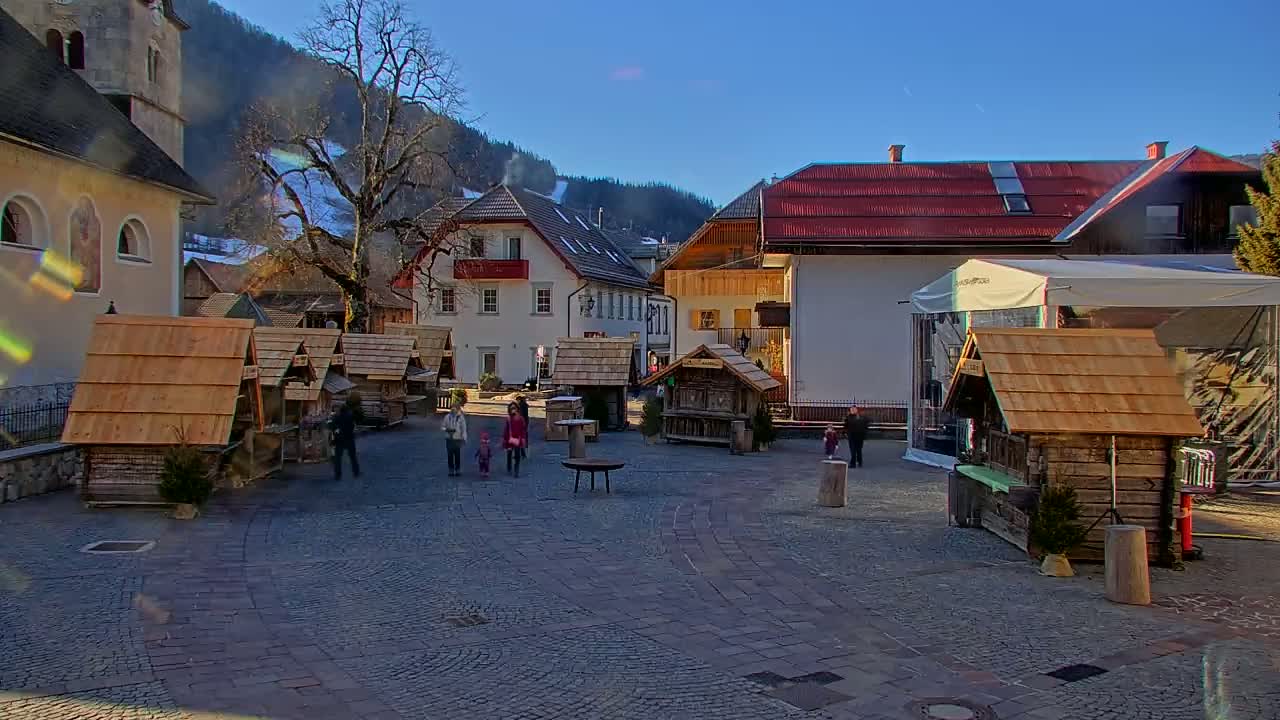 A cobblestone town square in a mountainous area features several small wooden stalls, a few people, and buildings with red-tiled roofs under a sunny sky.