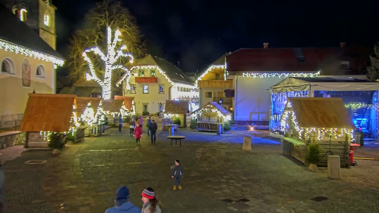 A nighttime scene of a town square decorated with numerous small wooden huts adorned with string lights, a large tree also lit with lights, and a stage set up for a performance.