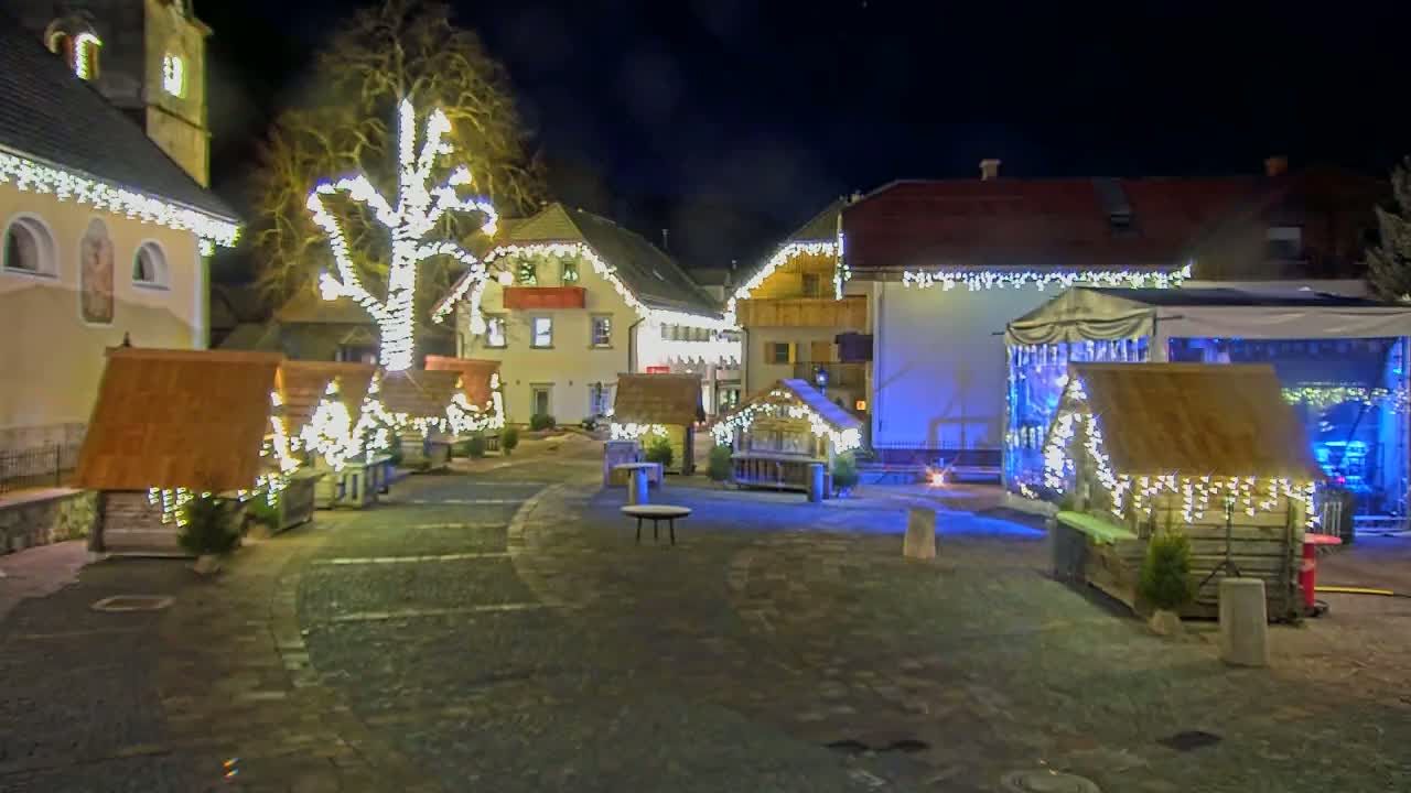A village square at night is decorated with string lights on buildings and small wooden structures, illuminated by numerous lights.