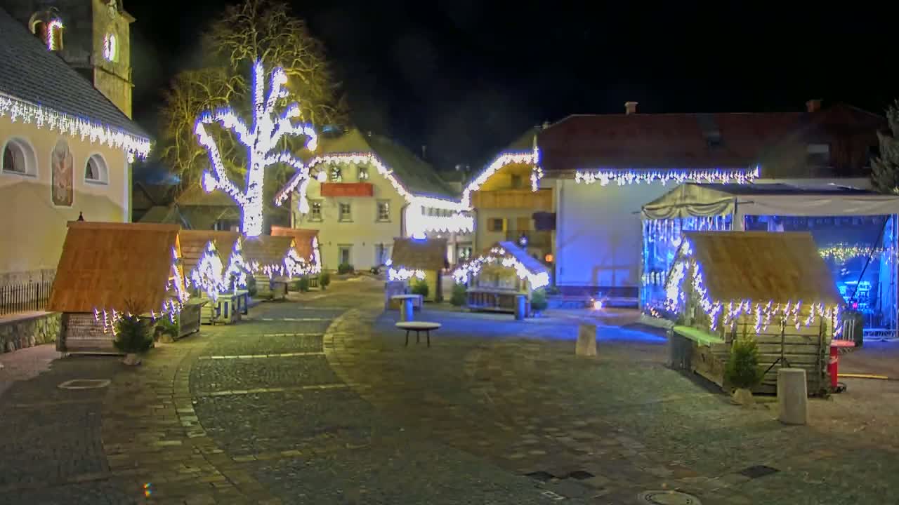 A nighttime town square is decorated with small wooden huts, string lights, and a large tree, all illuminated against a dark sky.