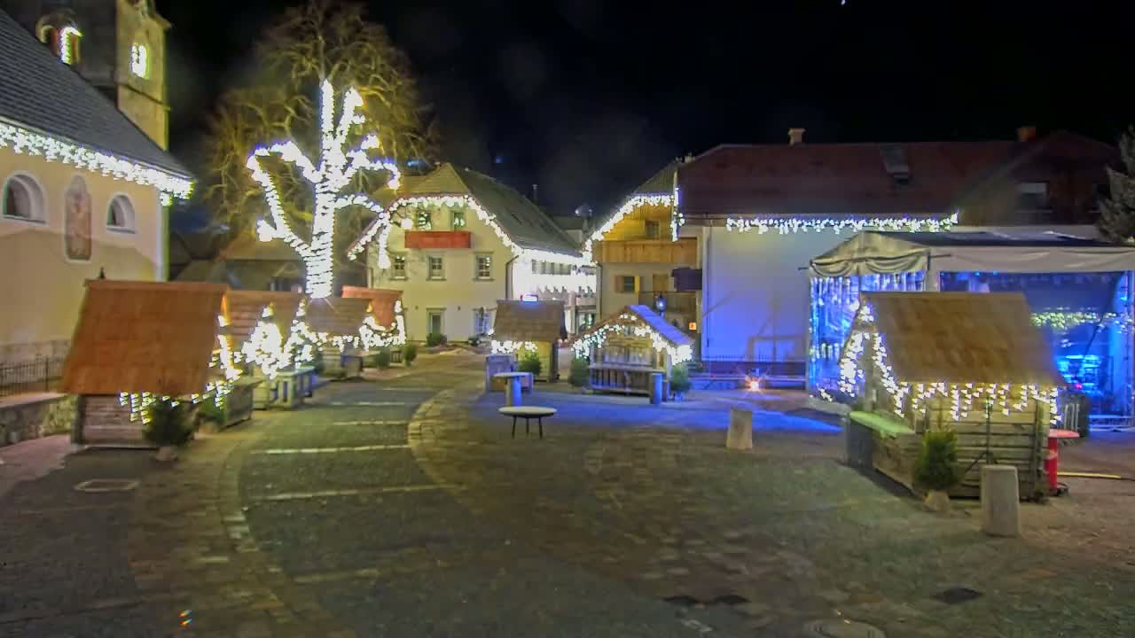 A nighttime scene shows a town square decorated with small wooden huts, a large tree, and strings of lights illuminating the buildings and cobblestone streets.