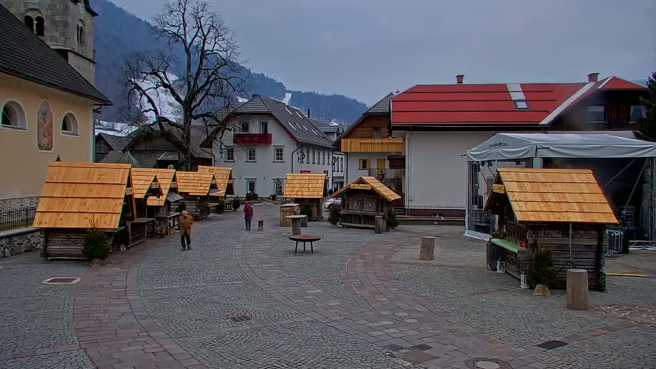 A cobblestone town square, under an overcast sky, features several small wooden stalls, buildings with alpine-style architecture, and a person walking a dog.