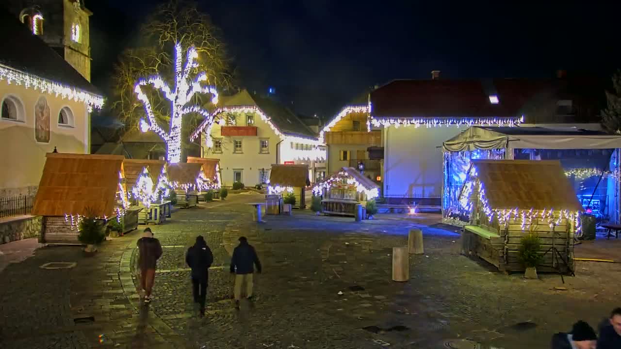 A nighttime scene of a town square decorated with Christmas lights and small wooden market stalls, with a few people walking through the cobblestone area.