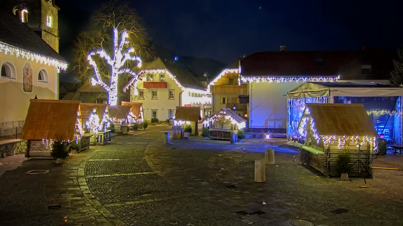 A village square at night is decorated with Christmas lights on buildings, trees, and small wooden stalls.