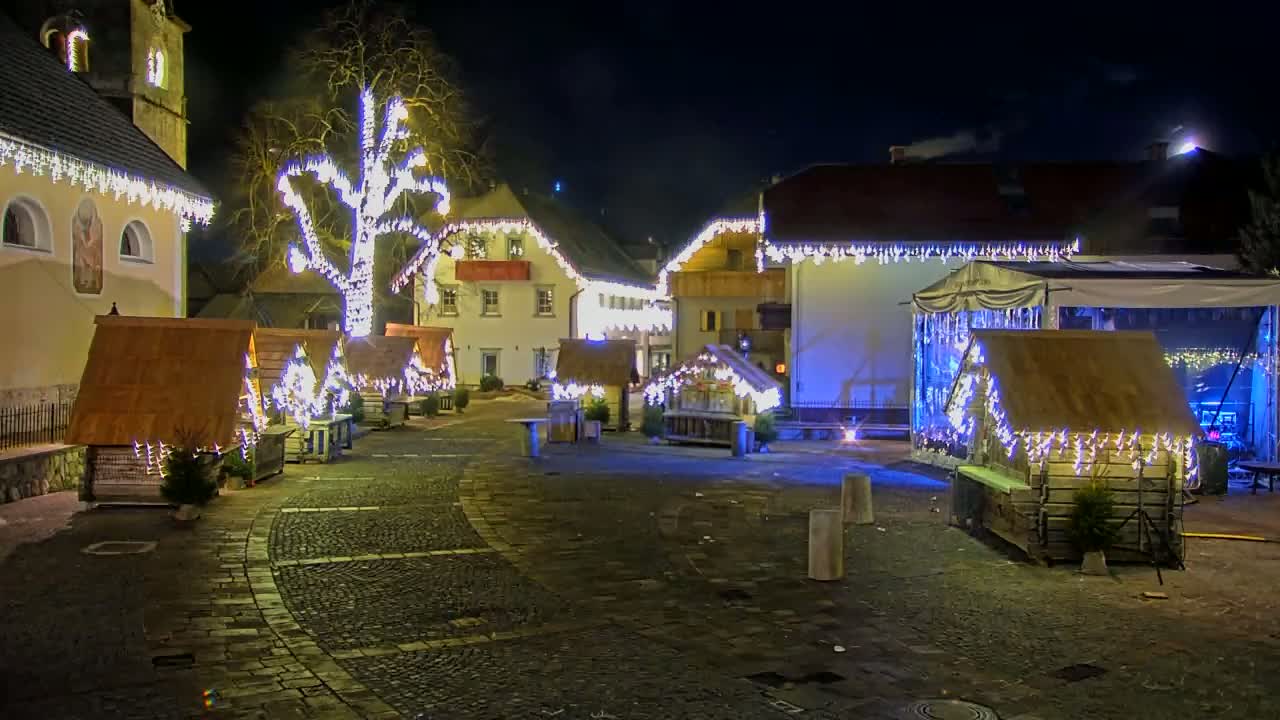 A nighttime scene of a town square decorated with Christmas lights and small wooden huts, under a clear, dark sky.