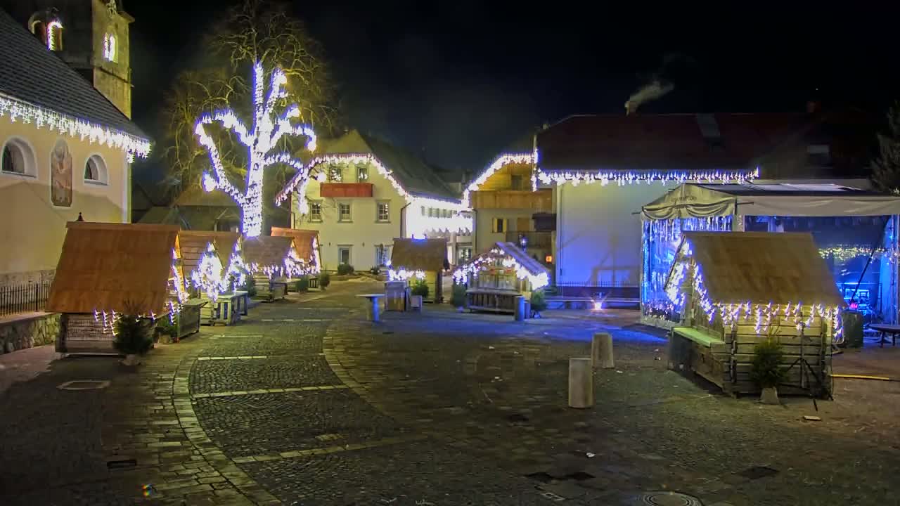 A cobblestone town square at night is illuminated by strings of lights adorning buildings and small wooden structures, with a large tree in the center also lit up.