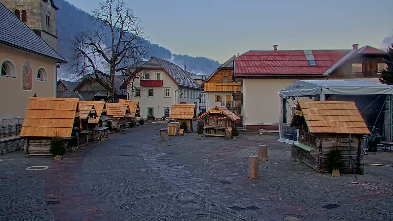 A cobblestone town square features several small wooden market stalls and buildings, with a backdrop of mountains under a mostly clear sky.