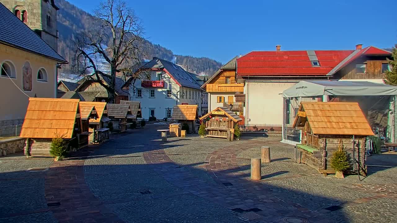 A cobblestone town square features several small wooden stalls, surrounded by buildings, under a clear sunny sky.