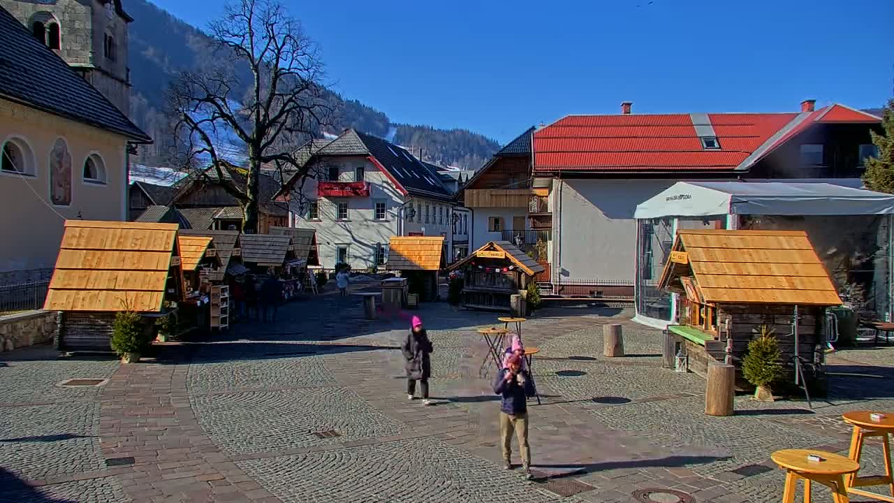 A cobblestone town square with several small wooden stalls, a few people, and buildings including a church, under a clear blue sky.