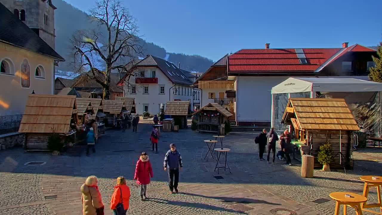 A sunny village square features several small wooden market stalls, surrounded by buildings and people walking on a cobblestone surface, with snow-covered mountains in the background.