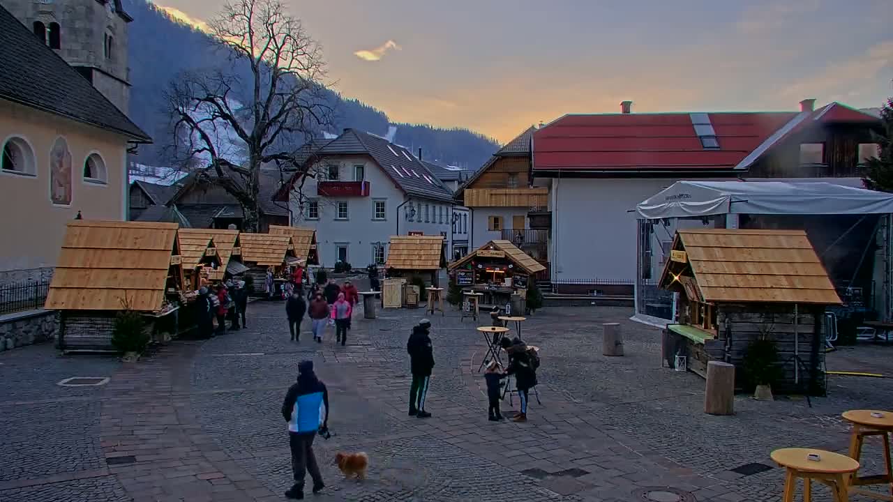 A town square with several wooden market stalls, people walking around, and a church and buildings in the background under a twilight sky.