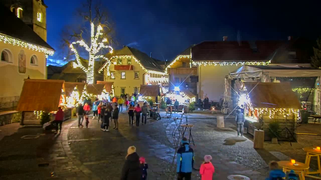 A nighttime outdoor Christmas market in a village square, illuminated by string lights and twinkling lights on wooden stalls and a large tree.