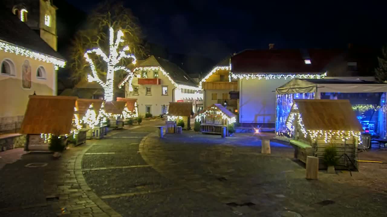 A nighttime scene of a town square decorated with Christmas lights illuminating small wooden huts and a large tree, under a dark sky.
