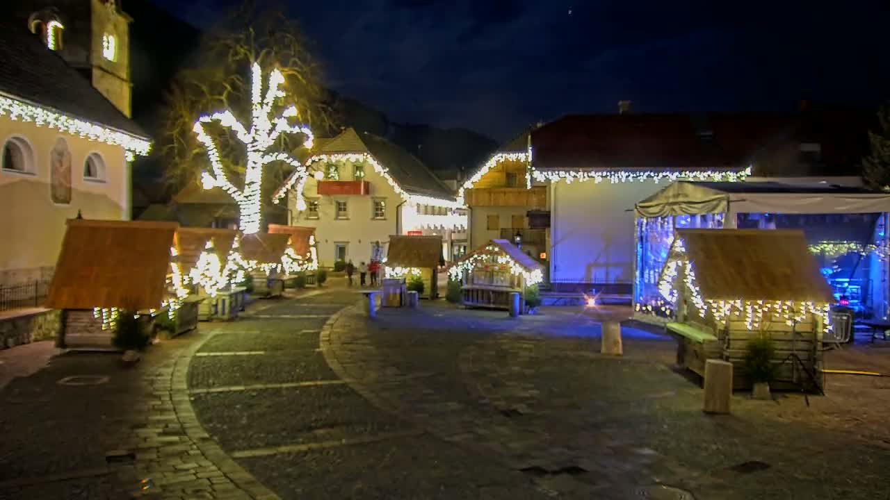 A nighttime scene of a village square decorated with Christmas lights adorning buildings and small wooden structures, with a large tree in the center also lit up.