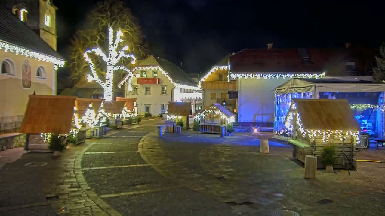 A town square at night is decorated with Christmas lights strung on buildings and small wooden stalls.