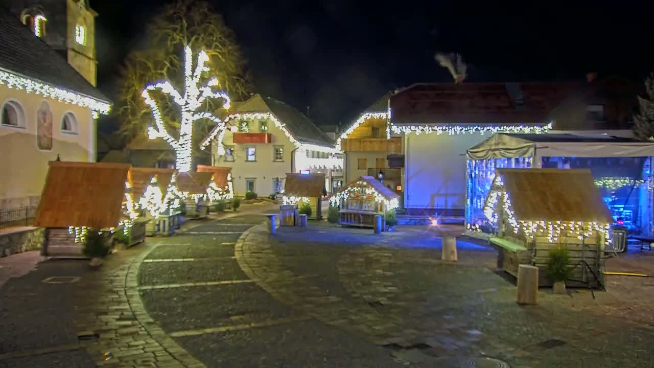 A town square at night is illuminated with strings of lights adorning buildings and small wooden structures, creating a festive atmosphere.