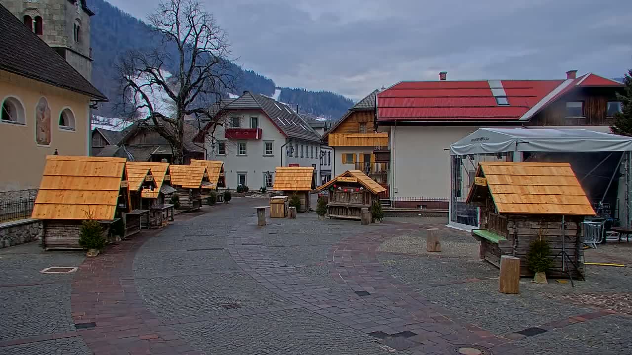 A cobblestone town square with several small wooden stalls, surrounded by buildings and a mountain backdrop under an overcast sky.