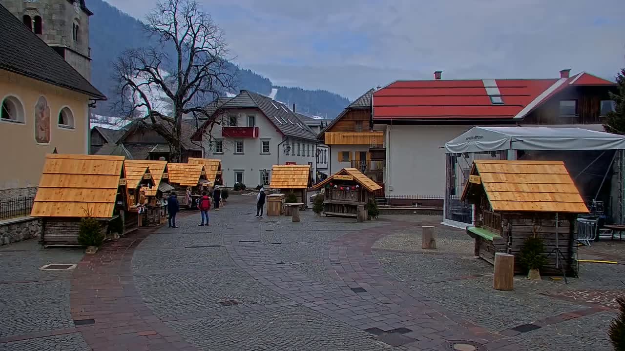 A town square with several small wooden market stalls, a church, and buildings in the background under an overcast sky.