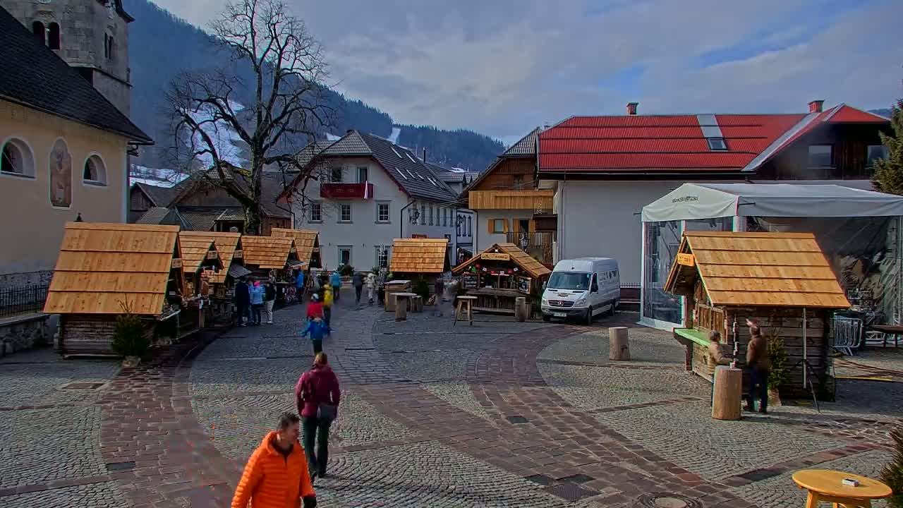 A cobblestone town square, under a partly cloudy sky, features several small wooden market stalls, a few pedestrians, and buildings with alpine-style architecture nestled against a snow-dusted mountainside.
