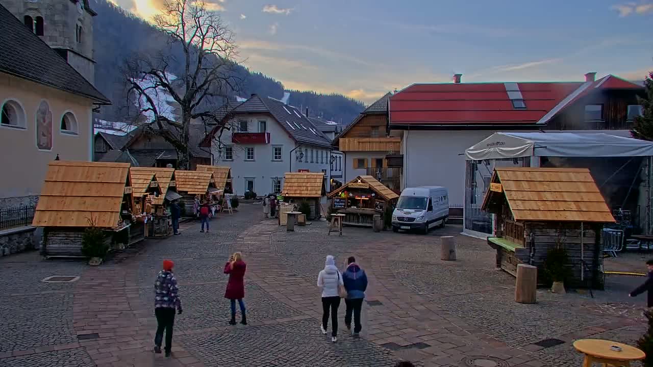 A cobblestone town square with several wooden market stalls, surrounded by buildings and a snow-covered mountain in the background under a partly cloudy sky.