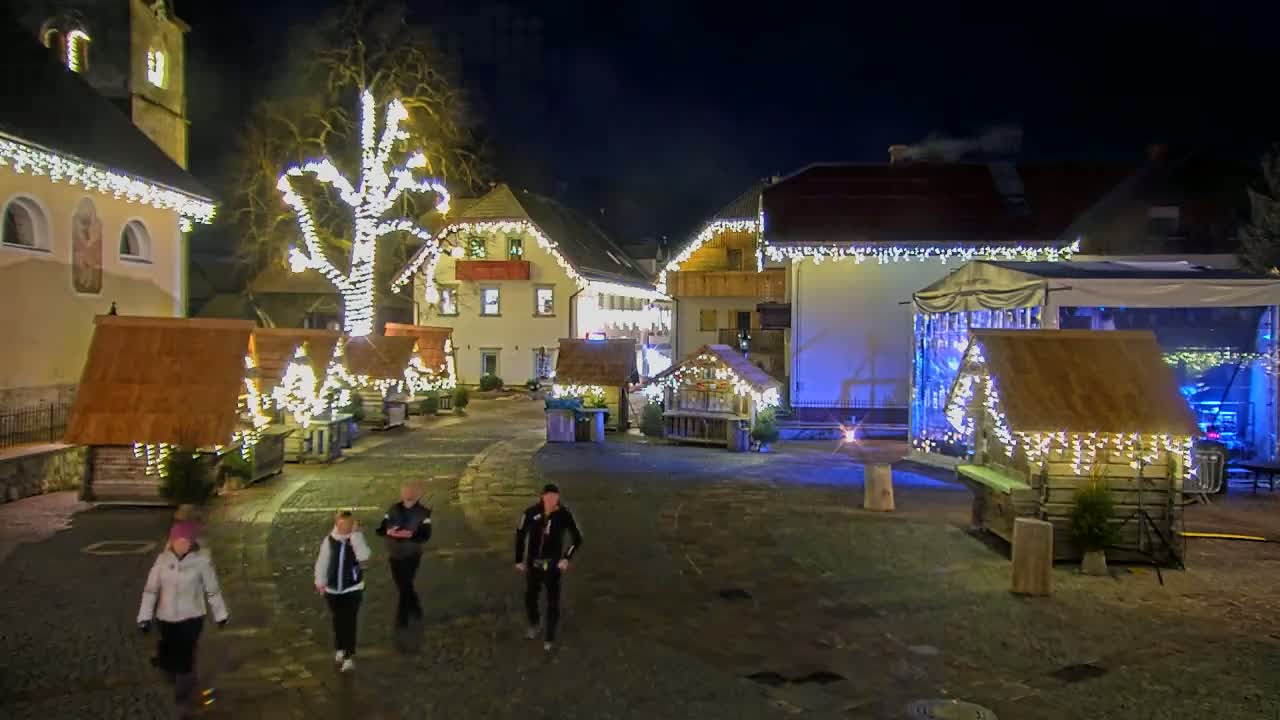 A nighttime view of a town square adorned with Christmas lights and small wooden stalls, with several people walking through the square.
