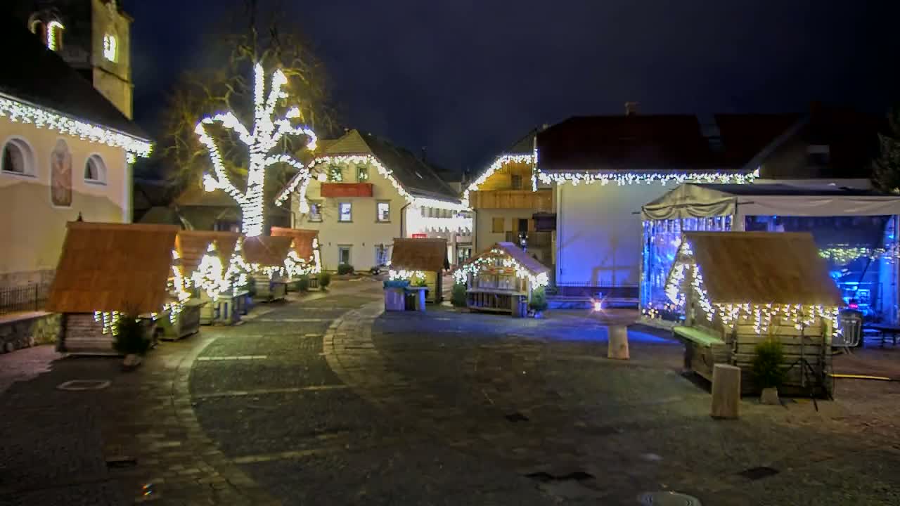 A nighttime scene of a town square decorated with Christmas lights illuminating small wooden stalls, a large tree, and buildings.