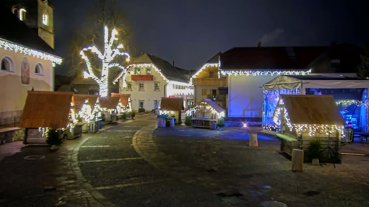 A nighttime outdoor scene shows a town square with small wooden market stalls and a large tree decorated with strings of white lights, all illuminated against a dark sky.