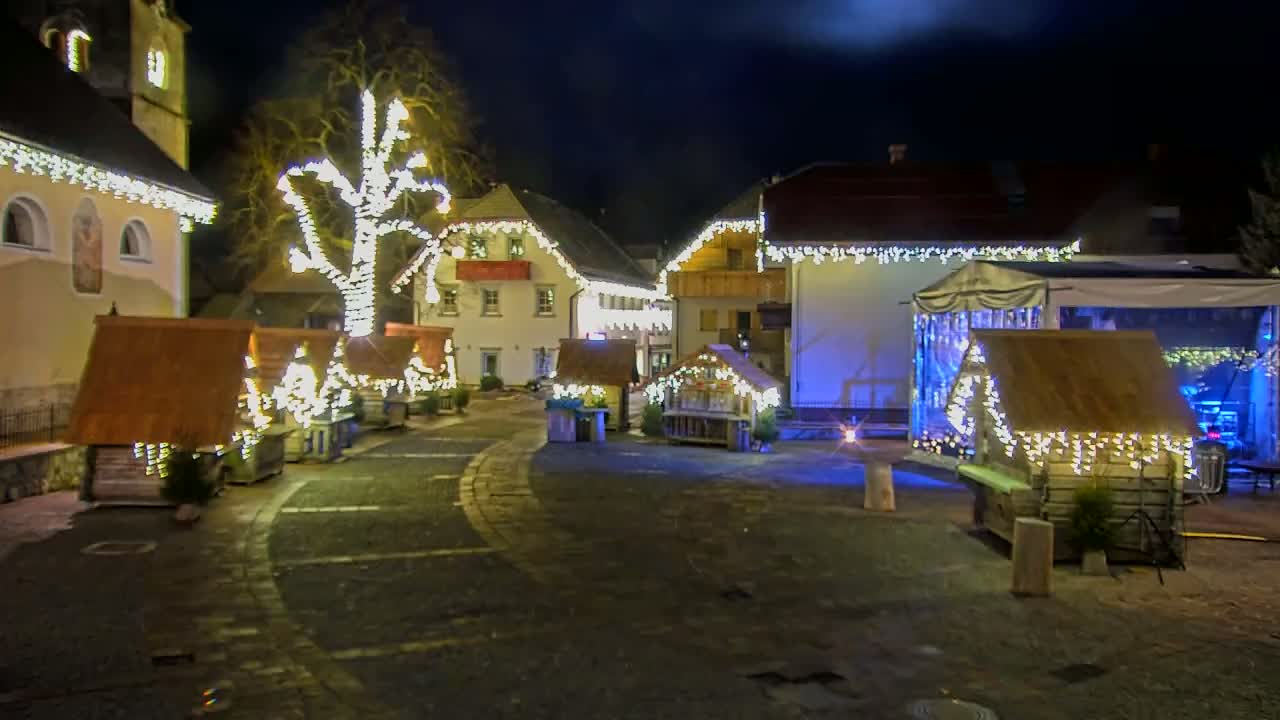 A nighttime village square is decorated with Christmas lights strung on buildings and small wooden stalls, illuminated by the lights and a partly cloudy night sky.