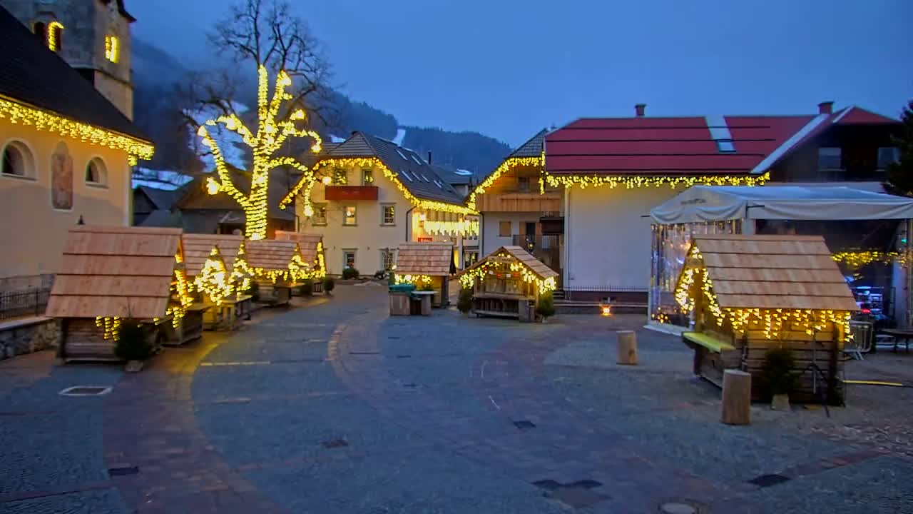 A town square at dusk is illuminated by string lights draped on buildings and small wooden market stalls, with a large tree also adorned with lights in the center.