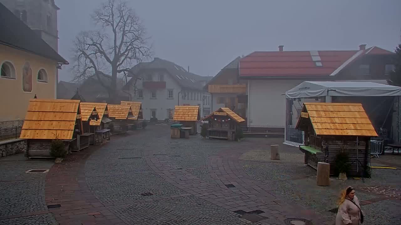 A foggy town square features several small wooden market stalls, a larger building with a red roof, and a church in the background.
