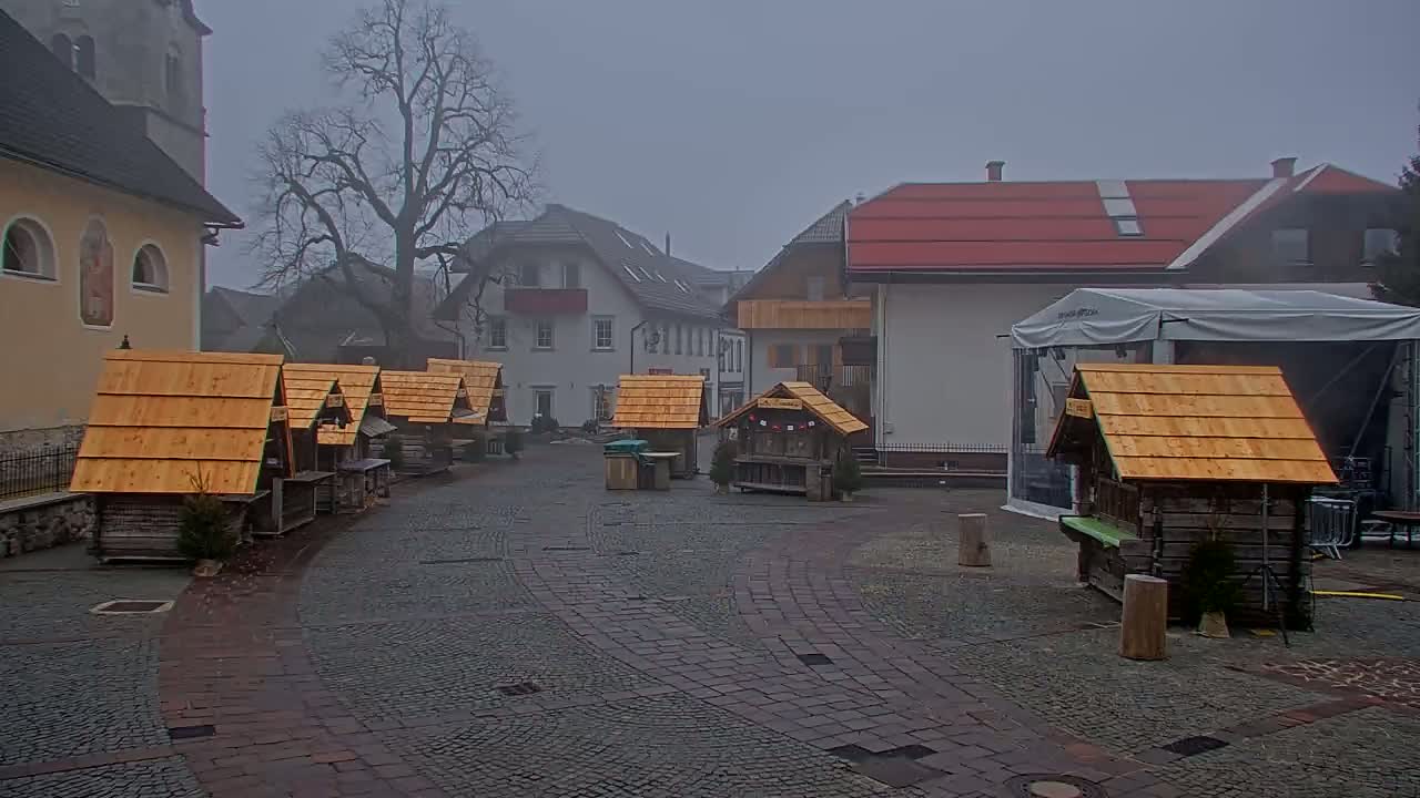 Several small wooden stalls with orange roofs sit in a foggy town square near buildings, one of which is a church.