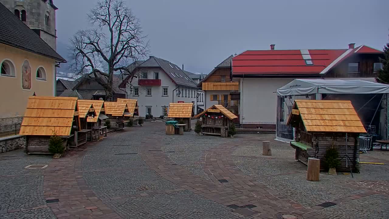 A stone plaza with several small wooden market stalls, surrounded by buildings on an overcast day.