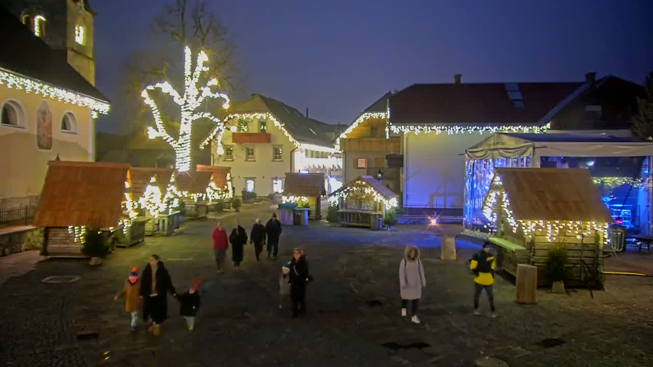 A nighttime outdoor Christmas market scene features several small wooden huts decorated with lights, a large tree also adorned with lights, and people strolling through the square.