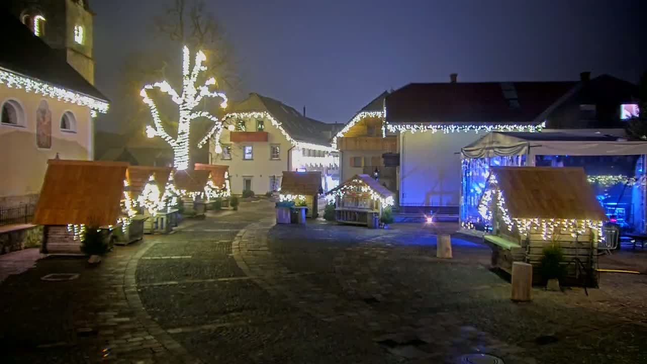 A nighttime village square is decorated with twinkling lights on buildings and small wooden stalls, and a tree is illuminated with lights.