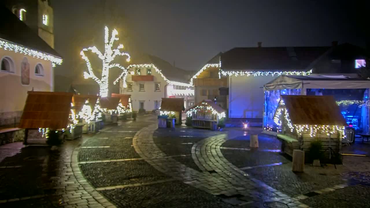 A cobblestone town square at night is decorated with numerous small wooden huts and a large tree, all illuminated by strings of warm white lights in foggy conditions.