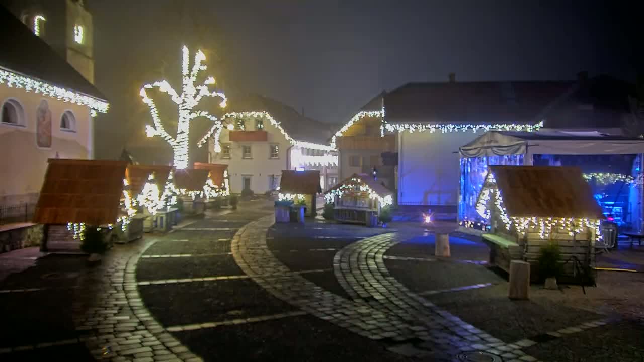 A foggy, nighttime scene of a town square decorated with Christmas lights adorning small wooden huts and a large tree, with buildings in the background also illuminated.