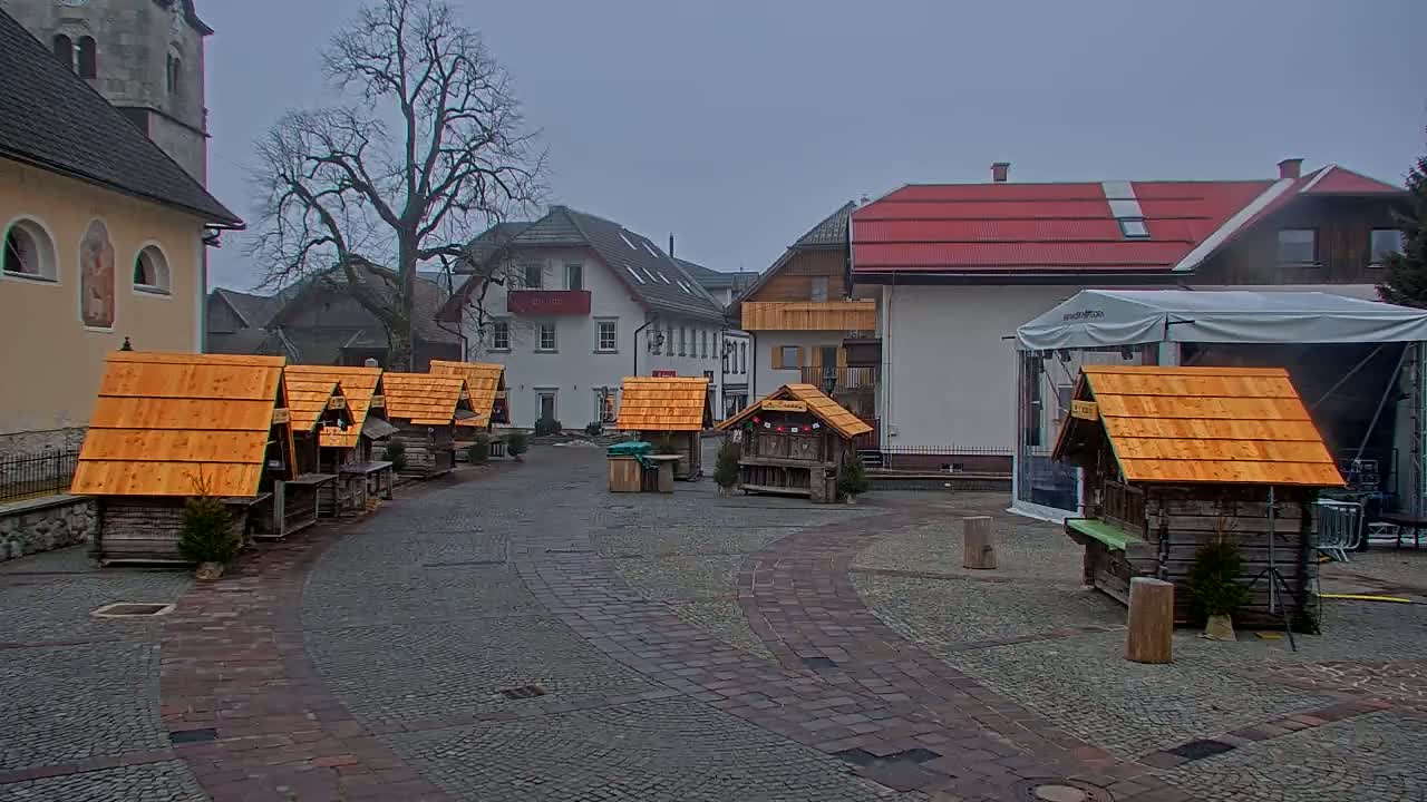 A town square on an overcast day features several small wooden stalls with orange roofs, situated near buildings and a large tent.