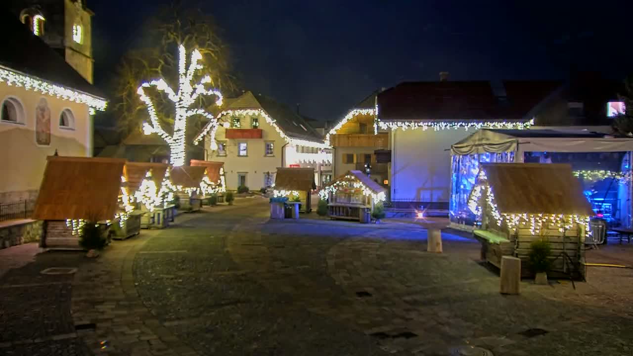 A nighttime scene of a town square with several small wooden huts decorated with string lights, a larger structure with a canopy, and buildings also adorned with lights.