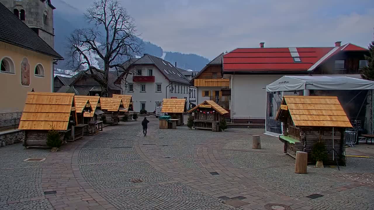 A cobblestone town square with several small wooden market stalls, a few buildings, and a person walking, all under an overcast sky.