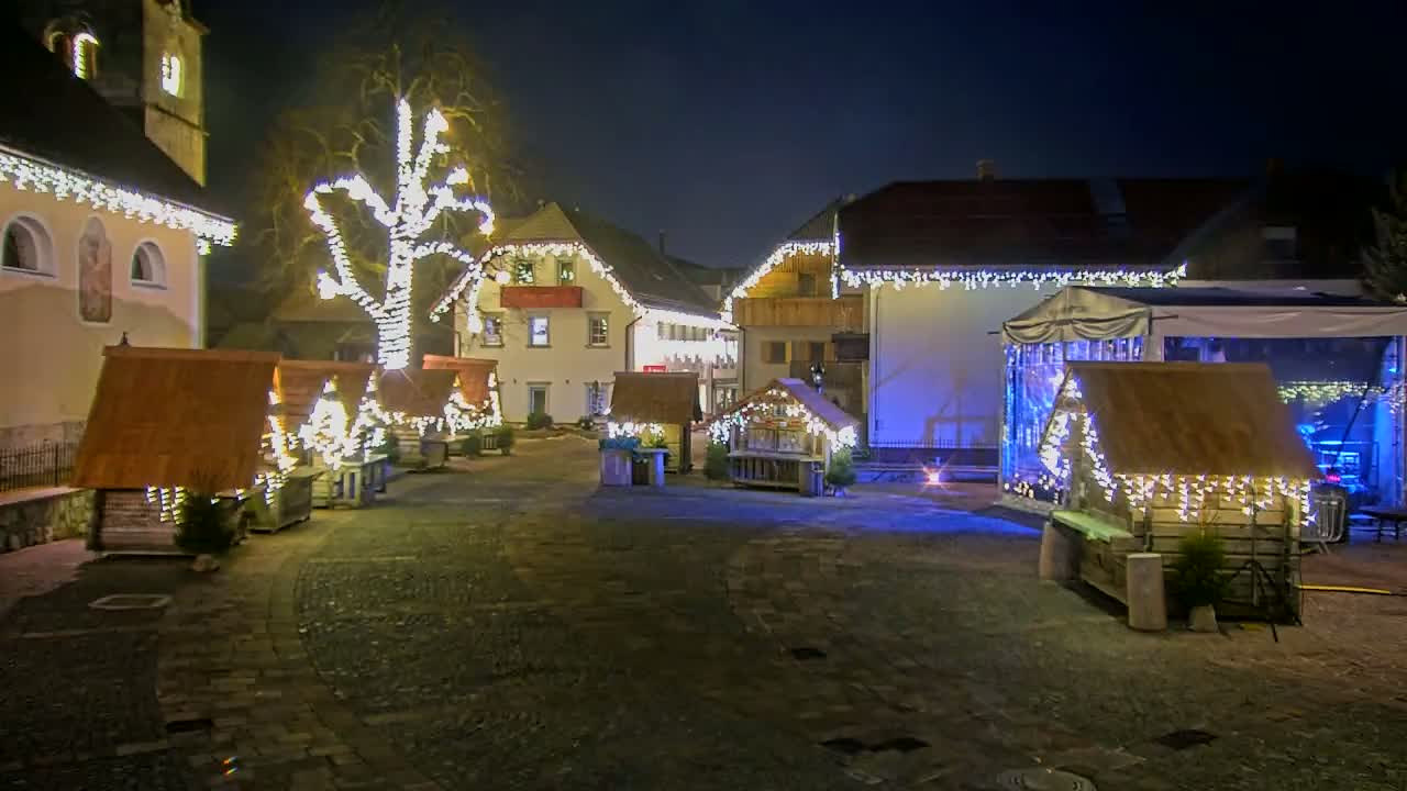 A nighttime scene of a town square with several small wooden huts decorated with string lights, a larger structure with a canopy, and a tree also adorned with lights.